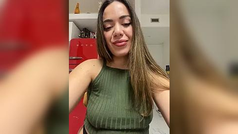 Media: Video of a young woman with long brown hair, wearing a green ribbed tank top, smiling in a kitchen with a red fridge, shelves, and a ceiling fan.