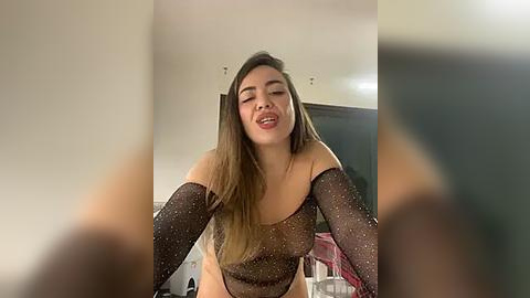 Media: Video of a young woman with long, straight, dark hair, wearing a sheer, black, sparkly top and matching long sleeves, leaning forward with a playful, open-mouthed smile. The background shows a dimly lit room with minimalistic decor.