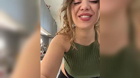 Media: A video of a smiling, curly-haired young woman in a green sleeveless top, standing in a modern kitchen with white cabinets and stainless steel appliances.