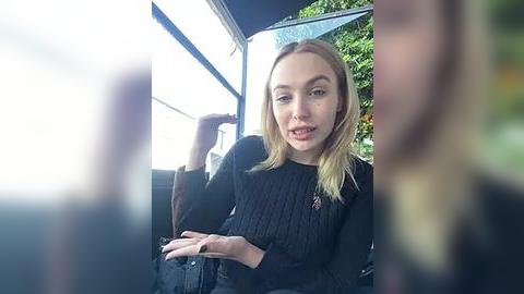 Media: Video of a young woman with fair skin and blonde hair, wearing a black sweater, seated in a bus, looking relaxed. Background shows blurred foliage and a window.