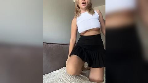 Media: Video of a blonde woman in a white crop top and black pleated skirt, kneeling on a textured white blanket, indoors with muted background.