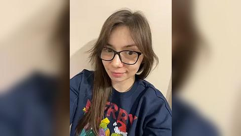 Media: Video of a young woman with long brown hair, wearing black-rimmed glasses and a dark blue \"Retro\" t-shirt, smiling, against a plain beige background.