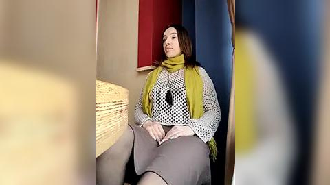 Media: Video of a woman with straight brown hair, wearing a yellow scarf and white patterned shirt, seated on a wicker bench against red and blue walls.