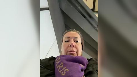 Media: A video of a middle-aged woman with light skin, short brown hair, and a serious expression, wearing a purple sweater and black jacket. She is standing inside a sleek, modern building with white tiles and dark metal railings.