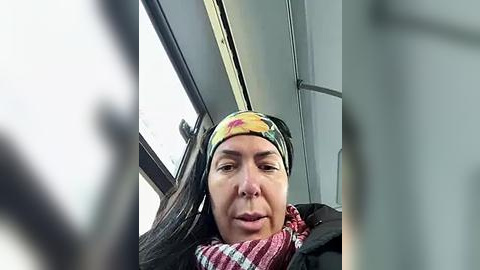 Media: Video of a woman with light skin, wearing a colorful headband, black jacket, and red scarf, sitting in a car with a window view of a snowy landscape.