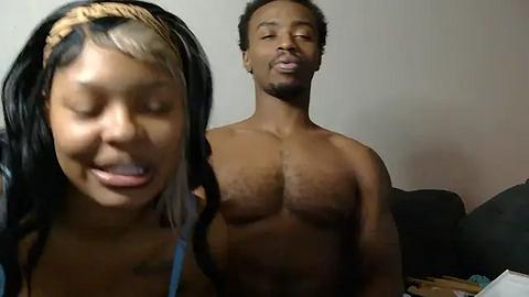 Media: Video of a topless African-American man with a muscular build and short hair, standing behind a smiling young African-American girl with straightened hair and a headband. They are indoors, on a dark couch against a plain background.