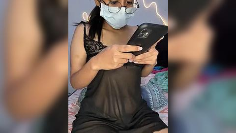 Media: Video of a woman in a sheer black lingerie top, wearing a face mask, reading a smartphone, with a blurred background featuring a bed and colorful blankets.