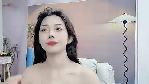 Media: A video of an East Asian woman with long black hair, light skin, and red lipstick, sitting topless on a white couch, facing a large, colorful beach scene mural.