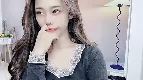 Media: Video of a young Asian woman with long, wavy brown hair, fair skin, and light makeup. She's wearing a dark gray sweater with white lace trim, looking pensively into a mirror. Background features a room with a lamp, plants, and a shelf.