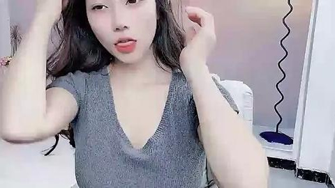 Media: Video of a young Asian woman with fair skin, long black hair, and a V-neck grey sweater. She is playfully touching her hair in a bathroom with a white sink and mirror.