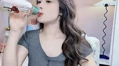 Media: A video of a young East Asian woman with long, wavy brown hair, wearing a grey short-sleeve shirt, drinking from a green and white plastic bottle in a brightly lit room with a desk, chair, and wall-mounted lamp.