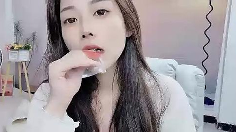 Media: A video of an East Asian woman with long black hair, wearing a white robe, and holding a clear plastic sheet to her lips, in a softly lit, minimalist room.