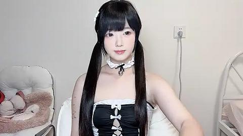 Media: Video of a young Asian woman with long black hair in pigtails, wearing a black and white maid outfit with a frilly collar and bow. She stands in a minimalist room with white walls and a small, white chair.