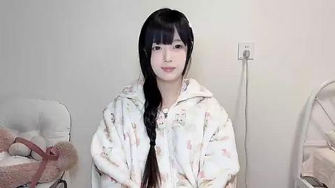 Media: Video of a young East Asian woman with long black hair in a white, floral-patterned robe, smiling gently. She stands against a plain, light-colored wall, with a stuffed animal and a mirror in the background.