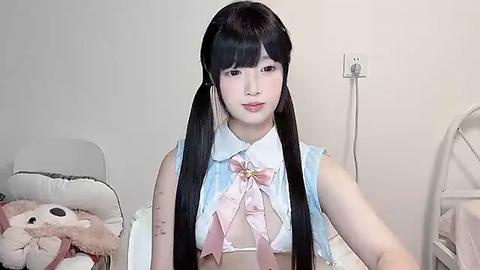 Media: Video of a young Asian woman with long black pigtails, wearing a white, frilly maid outfit with a pink bow, seated in a simple bedroom with beige walls and a fan.