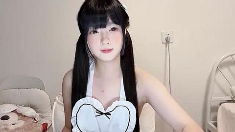 Media: Video of a young East Asian woman with long black hair and fair skin, wearing a white maid outfit with a heart-shaped neckline, sitting in a simple bedroom with a white bed and chair.