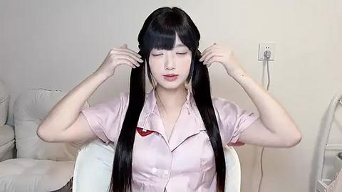 Media: Video of a fair-skinned East Asian woman with long black hair, wearing a pink blouse, adjusting her hair in a living room with white furniture and a fan.