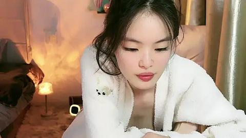 Media: Video of a young East Asian woman with fair skin, dark hair, and a petite frame, lying on a bed in a cozy room with warm, soft lighting. She wears a white robe, looking contemplative.