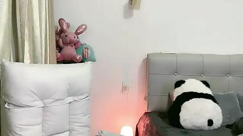 Media: A video of a baby's nursery with a pink bunny toy in a white crib, a tufted gray headboard, and a large black-and-white panda toy on a bed, lit by a soft, warm glow.