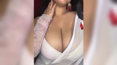 Media: Video of a light-skinned woman with large breasts, wearing a low-cut white dress with lace sleeves, red flower brooch, and red earrings. Her face is partially obscured by blurred figures in the background.