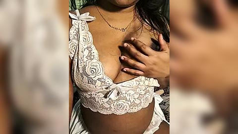 Media: Video of a young woman with medium brown skin, wearing a white lace bralette, holding her left breast, revealing a small nipple. Her long, dark hair is partially visible. Background is blurred.