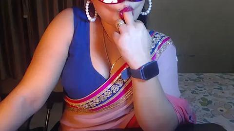 Media: Video of a woman with light skin, wearing a blue blouse, pink sari, pearl earrings, and a black smartwatch, biting her finger seductively. Background shows a bed with a floral sheet.