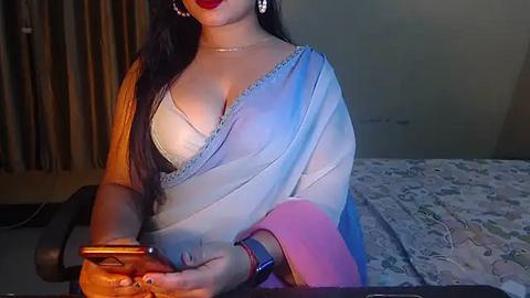 Media: A video of a woman with fair skin and long black hair, wearing a sheer blue sari with a white lace bralette, sitting on a floral bedspread, holding a smartphone, with a pink cushion beside her.