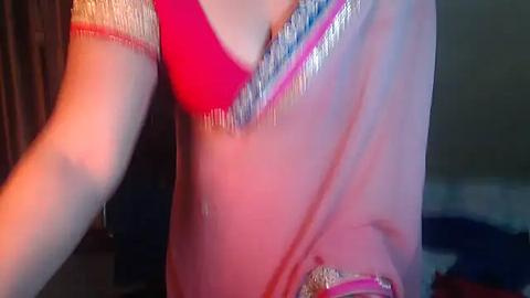 Media: Video of a person in a bright pink sari with golden embroidery, wearing a red blouse, taken indoors with dim lighting.