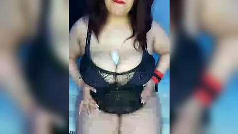 Media: Video of a plus-sized woman with fair skin and dark hair in a black, sheer lace lingerie bodysuit with a plunging neckline, sitting with hands on knees, wrists bound with red cuffs. Background is blurred.