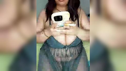 Media: A video shows a woman with medium brown skin, long dark hair, wearing a sheer, green, fringed top that reveals her breasts. She holds a white phone in front of her face, obscuring her upper body.