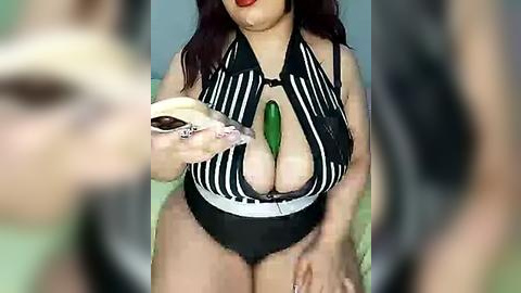 Media: Video of a woman with fair skin, wearing a black, striped, low-cut top with a large green gemstone, holding a cucumber.