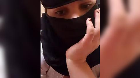 Media: A close-up video of a person with light skin, wearing a black face mask and black clothing, with their hand raised, partially obscuring their face.