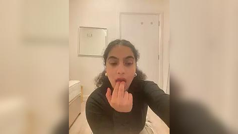 Media: A video of a young woman with curly hair, wearing a black top, licking her fingers, taken from a selfie angle in a beige hallway with a mirror and door.