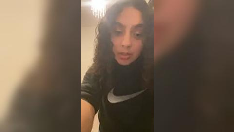 Media: Video of a young, curly-haired Latina woman with light skin, wearing a black t-shirt with a white Nike logo, looking downward, in a dimly lit room with blurred background.