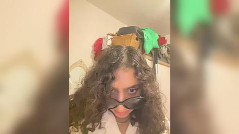 Media: Video of a person with curly black hair, wearing black sunglasses and a white shirt, balancing a stack of items including a green object, a black hat, and a red object on their head, in a dimly lit room.