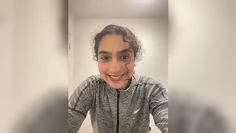 Media: Video of a young woman with curly dark hair, wearing a gray athletic jacket, smiling, standing in a white-walled hallway.