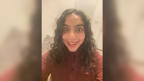 Media: Video of a young woman with curly black hair, wearing a maroon jacket, smiling broadly, taken from a slightly blurry selfie angle.