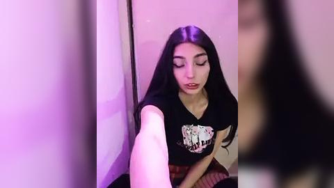 Media: Video of a young woman with long black hair, fair skin, and dark eyes, wearing a black graphic t-shirt, sitting against a light-colored wall. The image is blurred, with a purple tint and a slightly out-of-focus background.
