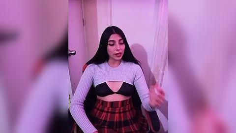 Media: Video of a young woman with long black hair, wearing a cropped light gray sweater and a red plaid skirt, sitting in a dimly lit room with a door and curtain visible in the background.