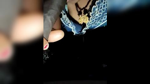 Media: A close-up video of a woman's hand in a black glove with red nail polish, adorned with a gold pendant necklace, resting on a blue patterned fabric.