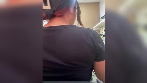 Media: Video of a woman's back in a supermarket, wearing a black T-shirt, her hair tied back, standing near a shelf. Background shows shelves and fluorescent lighting.