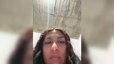 Media: A video of a Black woman with medium brown skin, wearing braided hair, looking serious, standing in front of a textured, light gray ceiling with a central spotlight.