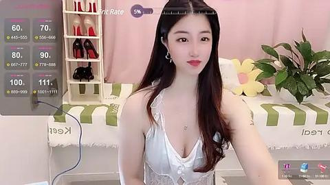 Media: Video of an East Asian woman with long dark hair, fair skin, and red lipstick, wearing a white lace bralette, sitting in a pastel room with a pink curtain, floral cushion, and shoes on a shelf.