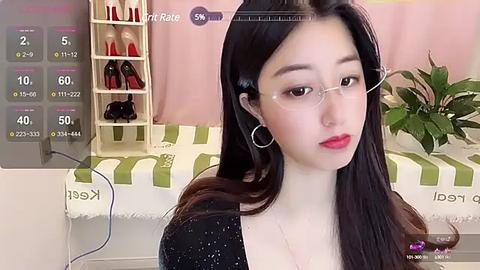 Media: Video of an East Asian woman with long black hair, fair skin, and wearing glasses, sitting in a room with a green-striped couch and a shelf of shoes.