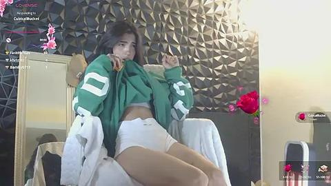 Media: Video of a young Asian woman with long black hair, wearing a green football jersey and white shorts, sitting on a bed with a geometric patterned wall behind her.