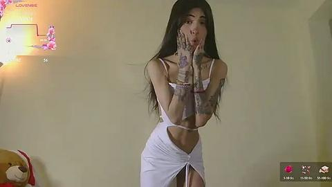 Media: A video of a slender woman with long black hair, wearing a white, sleeveless, high-waisted dress, with intricate tattoos covering her arms and hands. She stands in a dimly-lit room with a pink flower arrangement on the left.