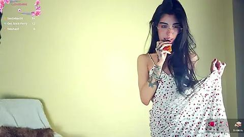 Media: Video of a slender, long-haired, dark-skinned woman in a white polka-dot dress, holding a glass of wine, in a brightly lit room with a green wall and a pink flower arrangement.