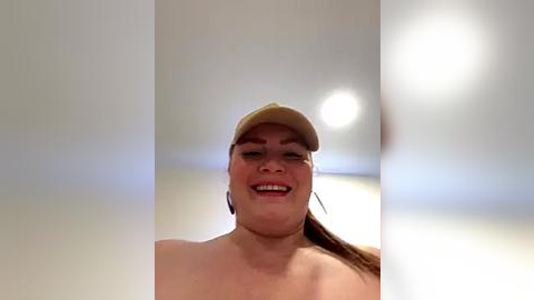 Media: Video of a smiling, topless woman with light skin, brown hair, and a beige baseball cap, standing against a blurred background with a bright light source.