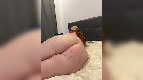 Media: Video of a nude, plus-sized woman with fair skin, freckles, and red hair, lying on her stomach on a bed with beige sheets. She's holding a smartphone, and a grey headboard is visible in the background.