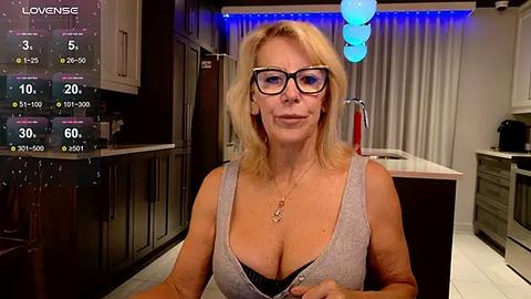 Media: Video of a blonde woman in glasses, wearing a low-cut gray tank top, sitting in a modern kitchen with blue lighting and a weather forecast overlay.